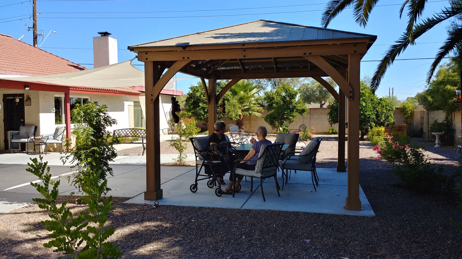 Elderly residents sit at a shaded wooden gazebo patio with chairs and a walker.