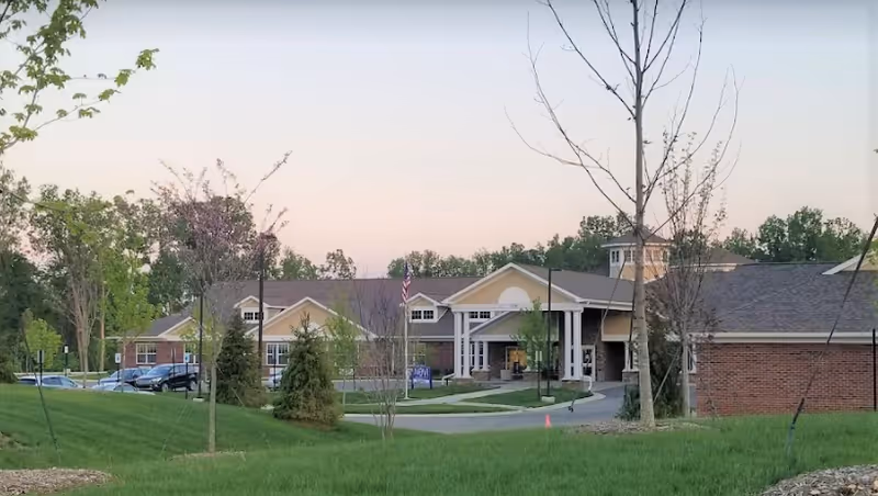 Exterior front view of Novi Lakes Health Campus building with a driveway, American flag, and landscaped lawn at dusk.
