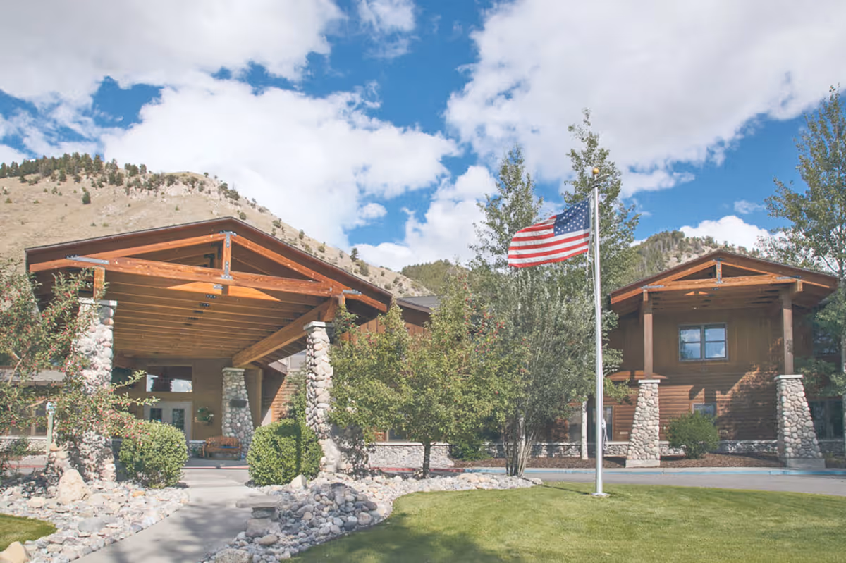 Exterior view of Legacy Lodge at Jackson Hole, showing a building with wooden beams and stone pillars under a partly cloudy sky. There is an American flag on a flagpole in front of the building, surrounded by trees, bushes, and a landscaped area with rocks and grass.
