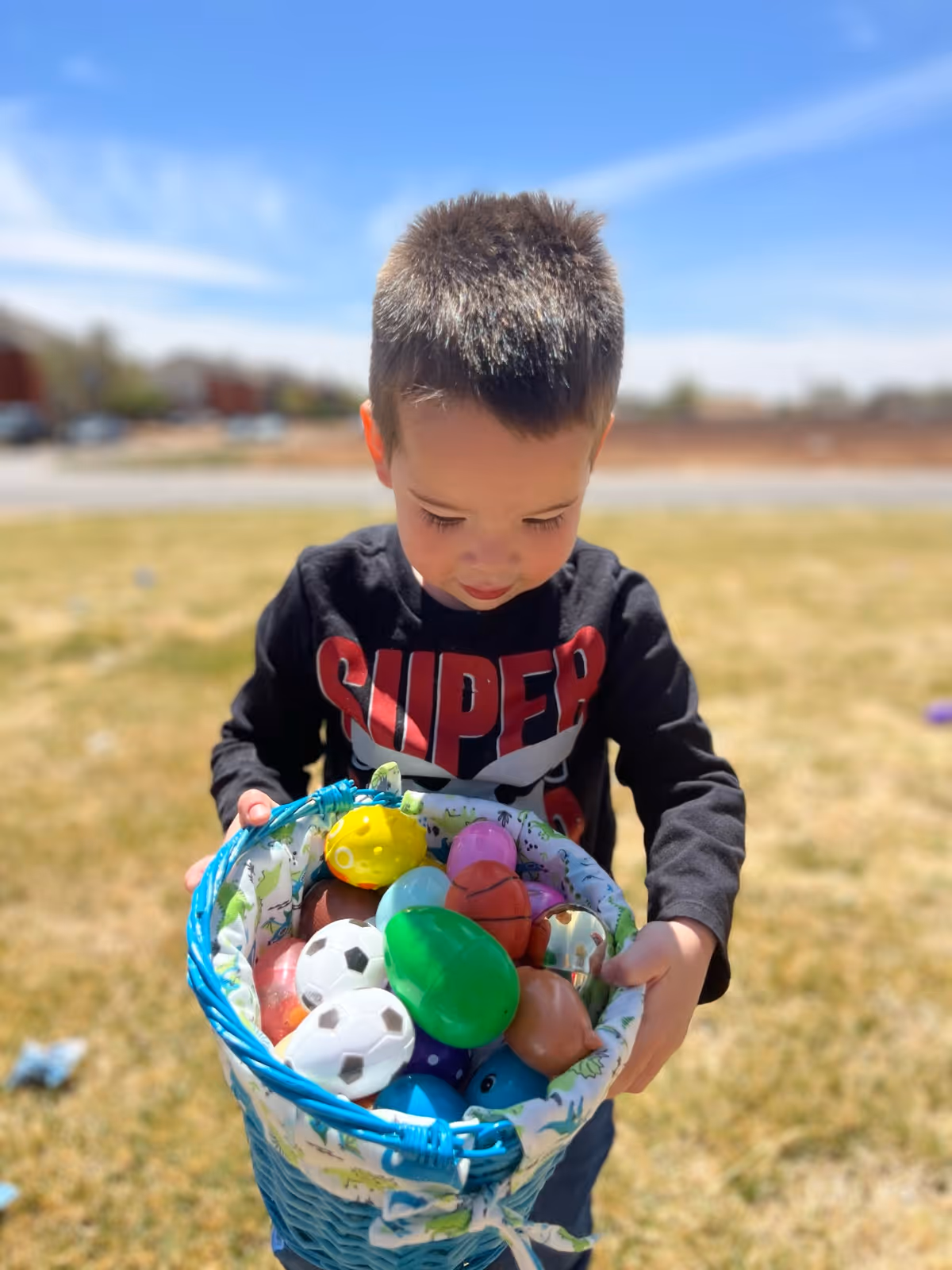 A young boy outdoors holding a blue basket filled with colorful plastic Easter eggs and small toy balls, standing on grass with a clear blue sky in the background.