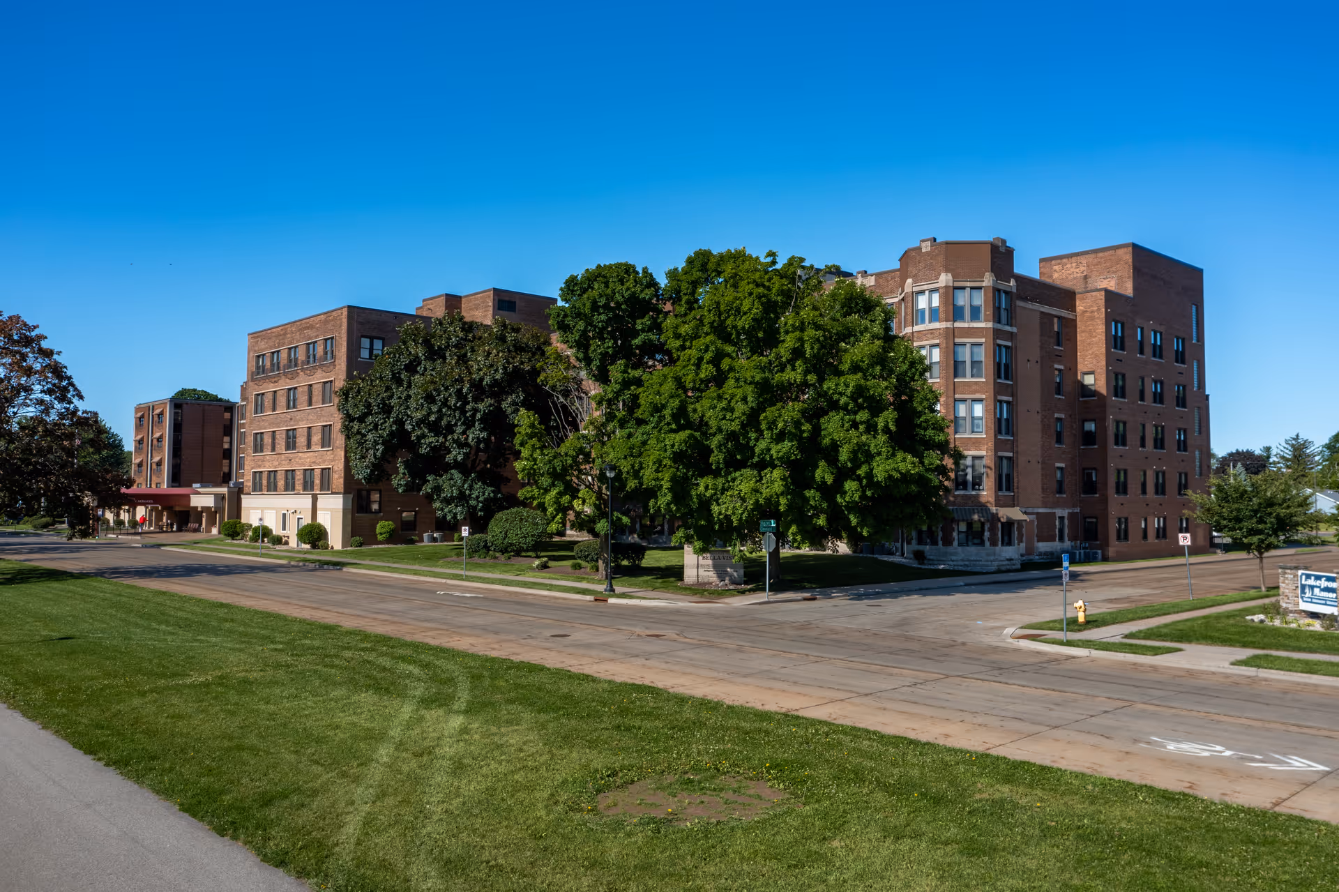 Exterior view of a large multi-story brick building with several windows, surrounded by green trees and grass under a clear blue sky. The building is located at a street corner with sidewalks and a road in front.