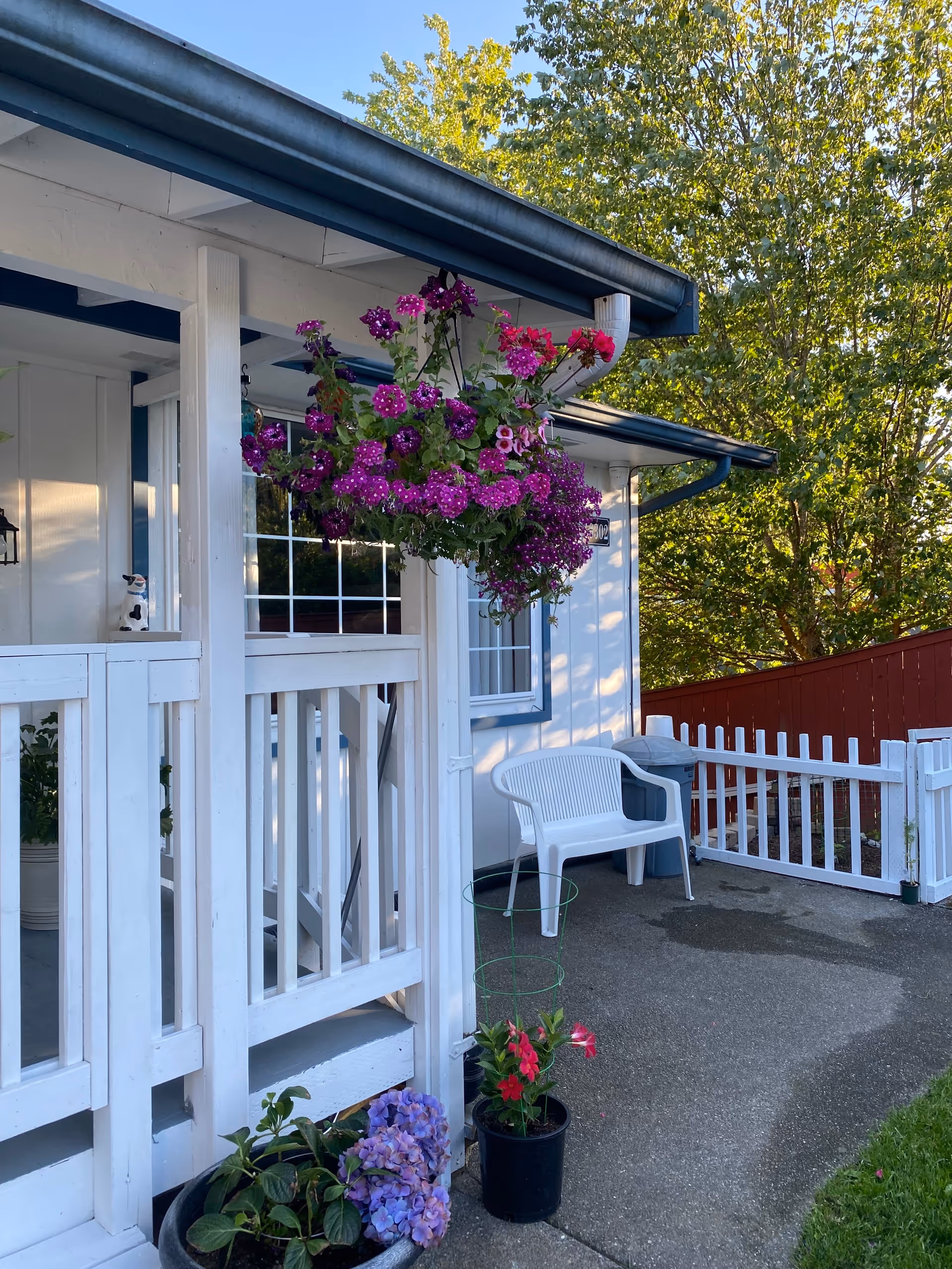 White porch with purple hanging flowers, potted plants, and a white plastic chair outside.