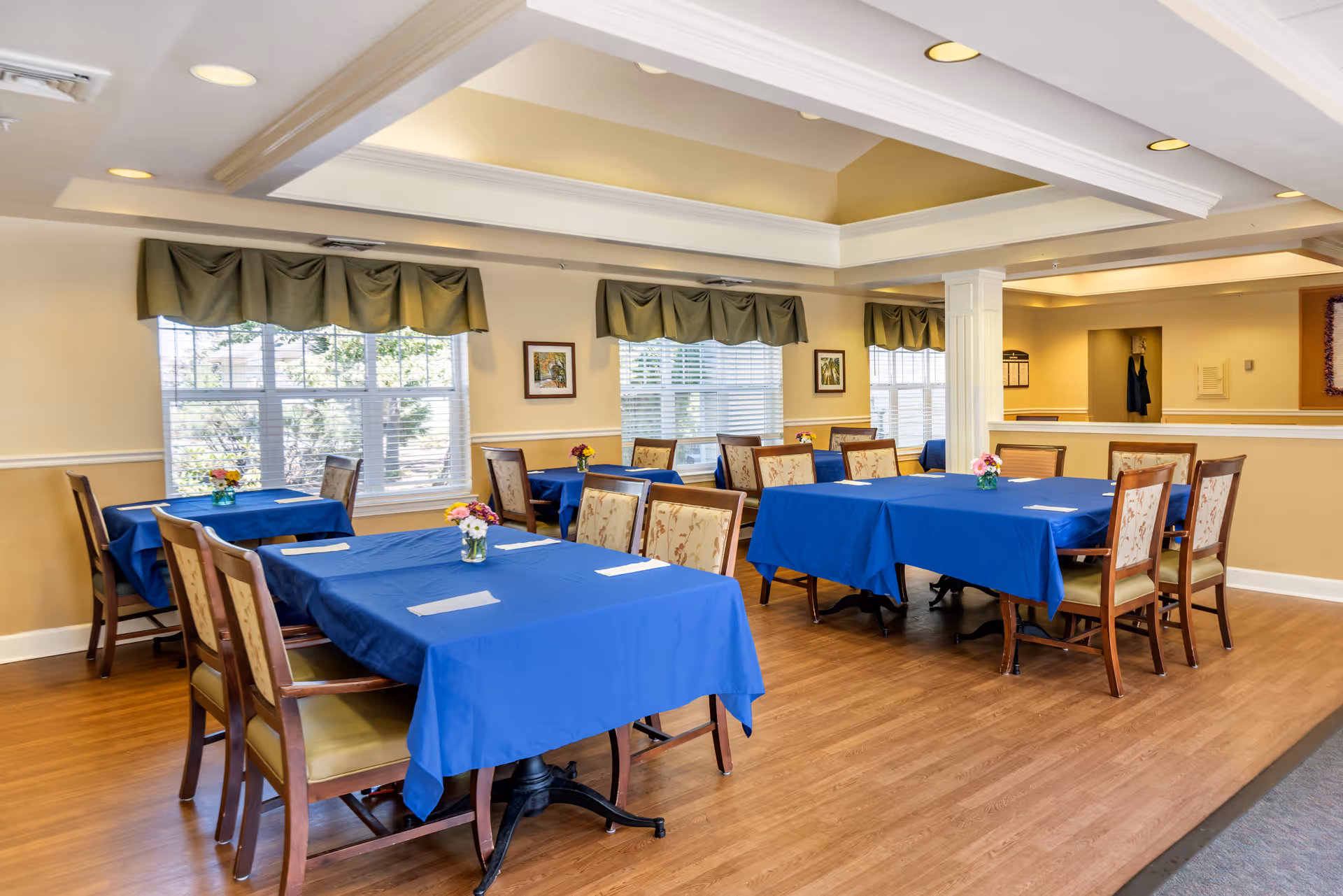 A bright dining room with several tables covered in blue tablecloths, each surrounded by wooden chairs with floral upholstery. Small flower arrangements are placed on each table. Large windows with green valances allow natural light to fill the room, and framed artwork hangs on the walls. The floor is wooden, and the ceiling features recessed lighting and a tray design.