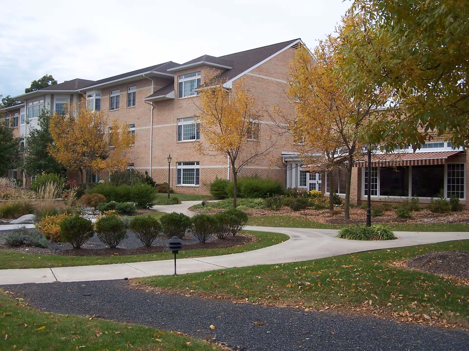 Exterior view of a three-story brick senior living facility with landscaped gardens, trees with autumn foliage, and paved walkways in the foreground.