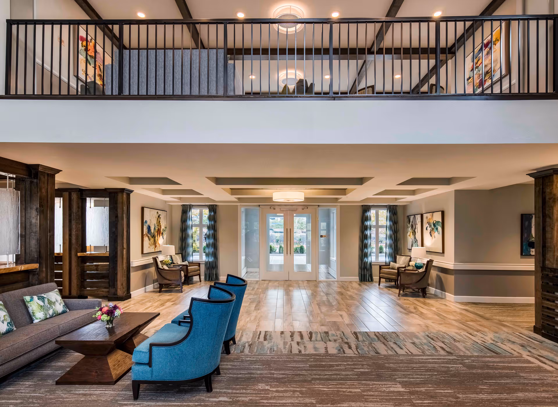 Spacious and well-lit senior living facility common area with a seating arrangement including a gray sofa, two blue armchairs, and a wooden coffee table with a flower vase. The room features wooden columns, large windows with patterned curtains, modern ceiling lights, and abstract artwork on the walls. A balcony with black railings overlooks the area.