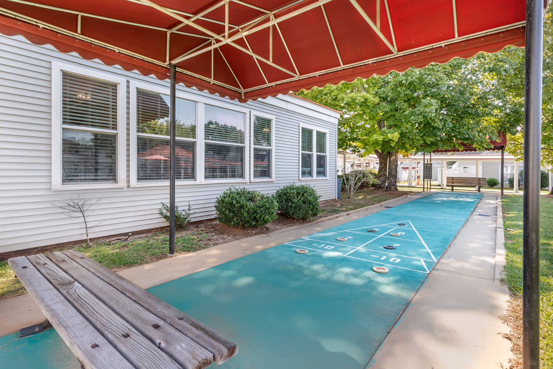 Outdoor shuffleboard court with a red canopy overhead, wooden benches on either side, and a white building with windows and bushes along the side. Trees and greenery are visible in the background.