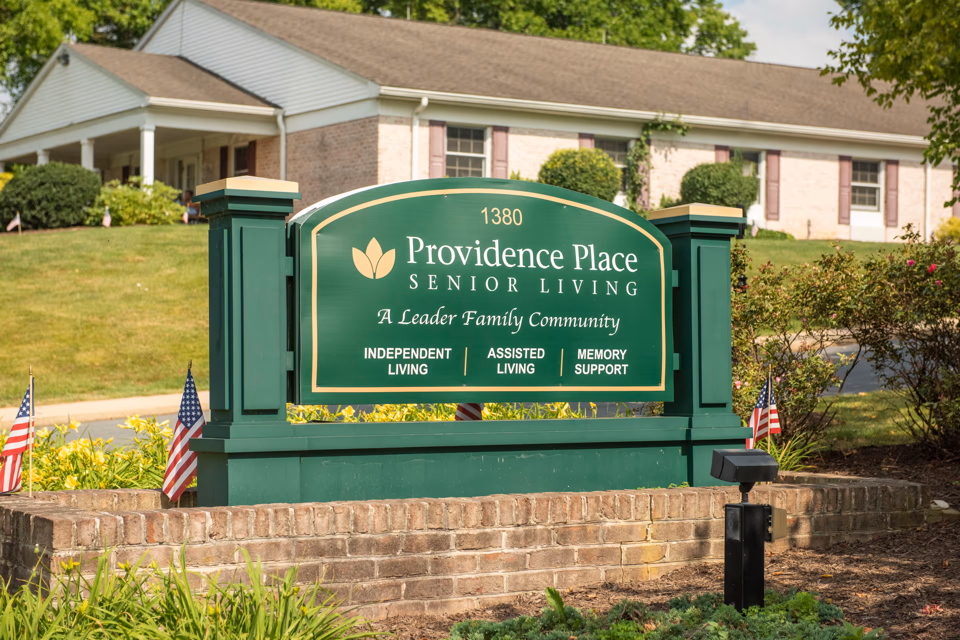 A green sign reading "Providence Place Senior Living" stands on a brick base in front of a single-story building with landscaping and small American flags.