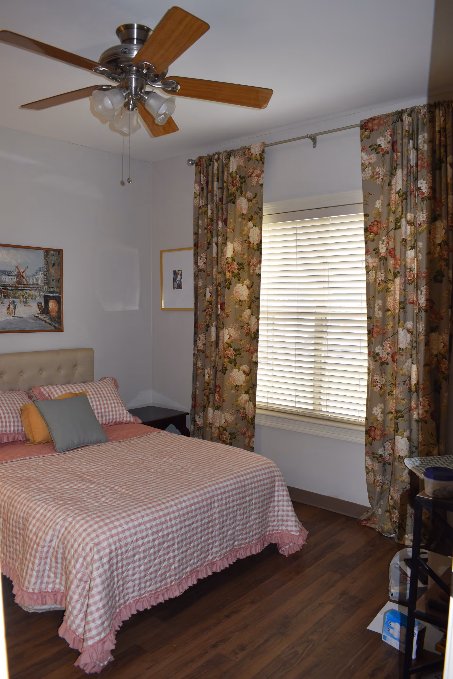 A bedroom in a senior living facility with a bed, floral curtains, and ceiling fan.