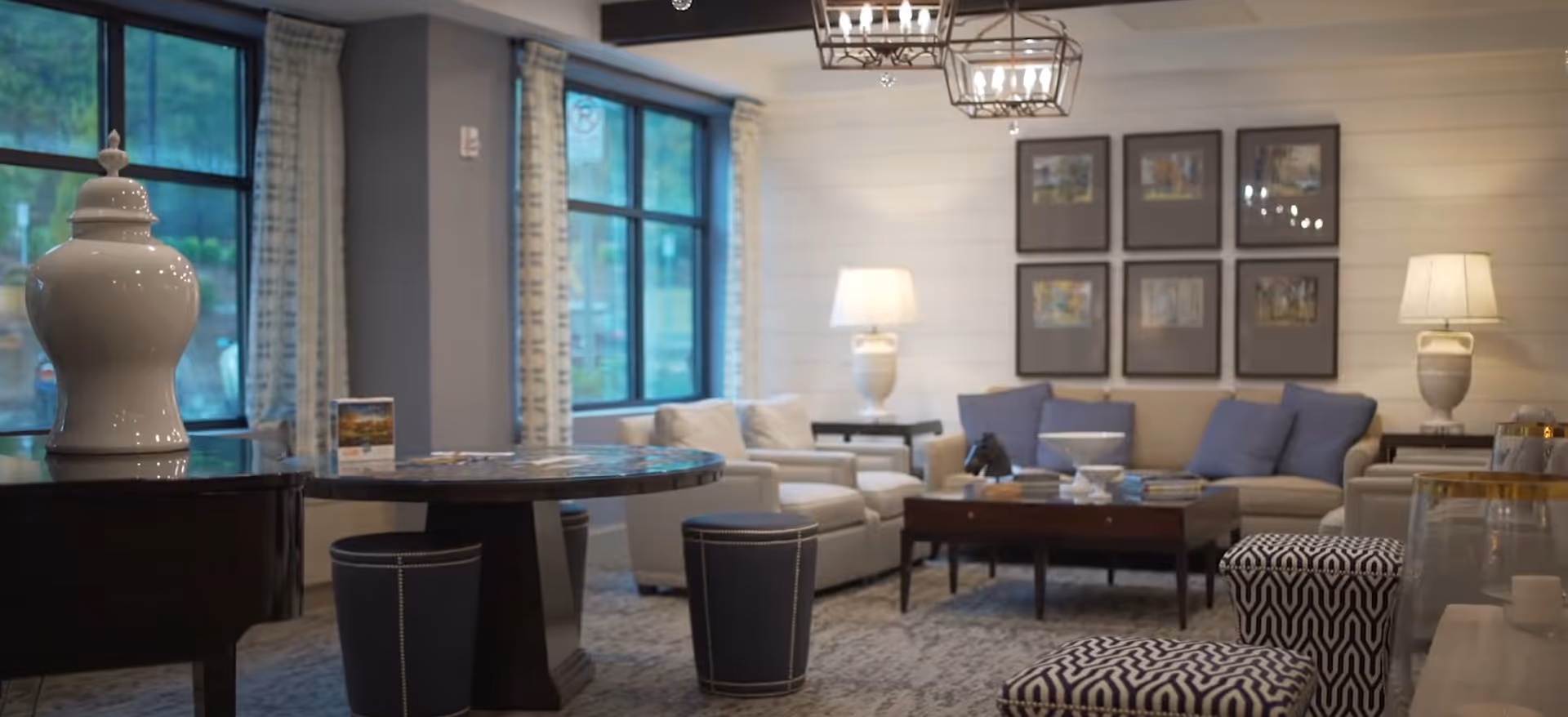 Well-lit communal living room with sofas, a coffee table, lamps, and patterned ottomans by large windows.