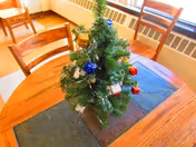 A small decorated tabletop Christmas tree sits on a wooden dining table with chairs by a window.