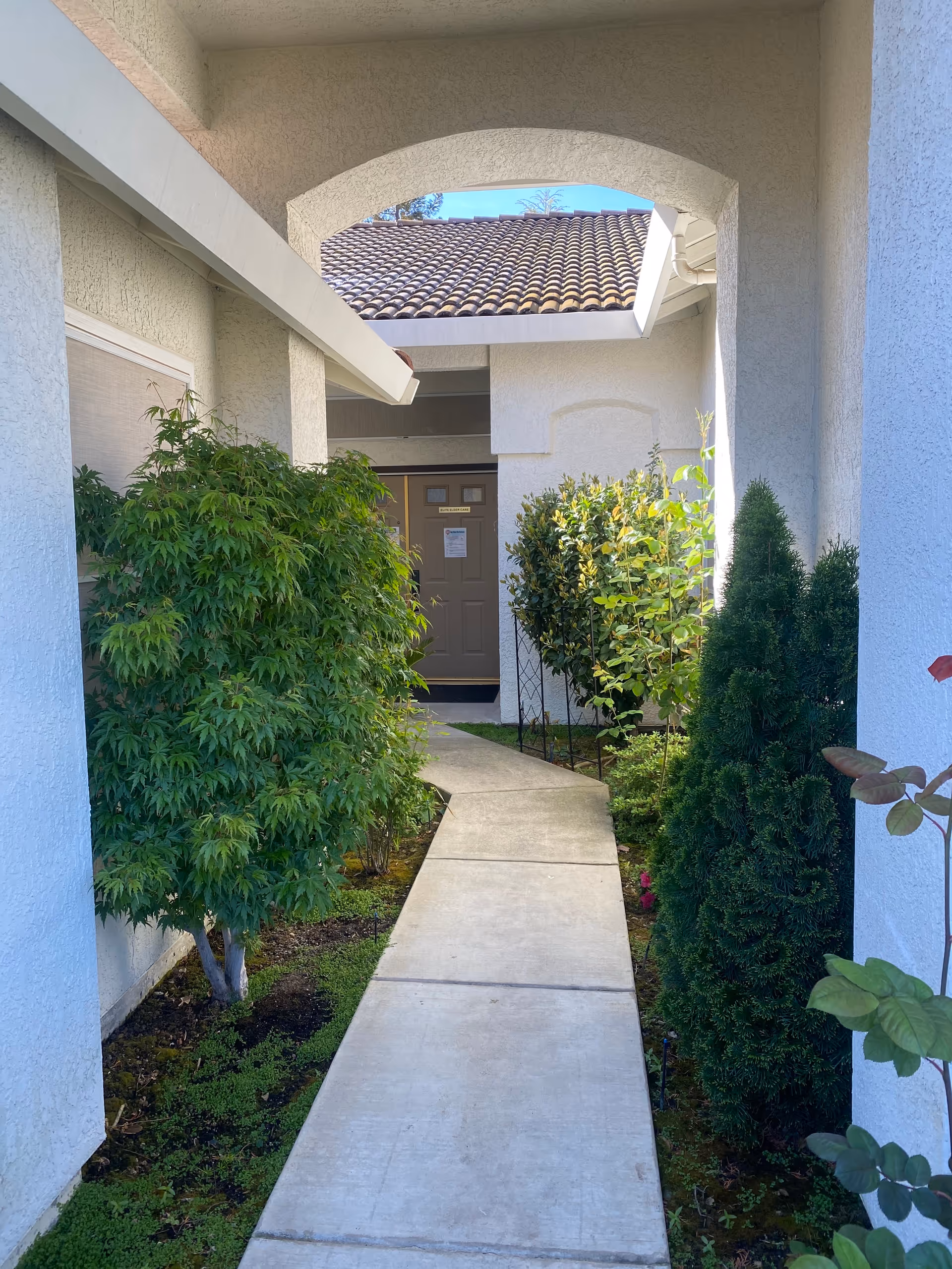Concrete walkway leads to a front door framed by green shrubs and an archway.