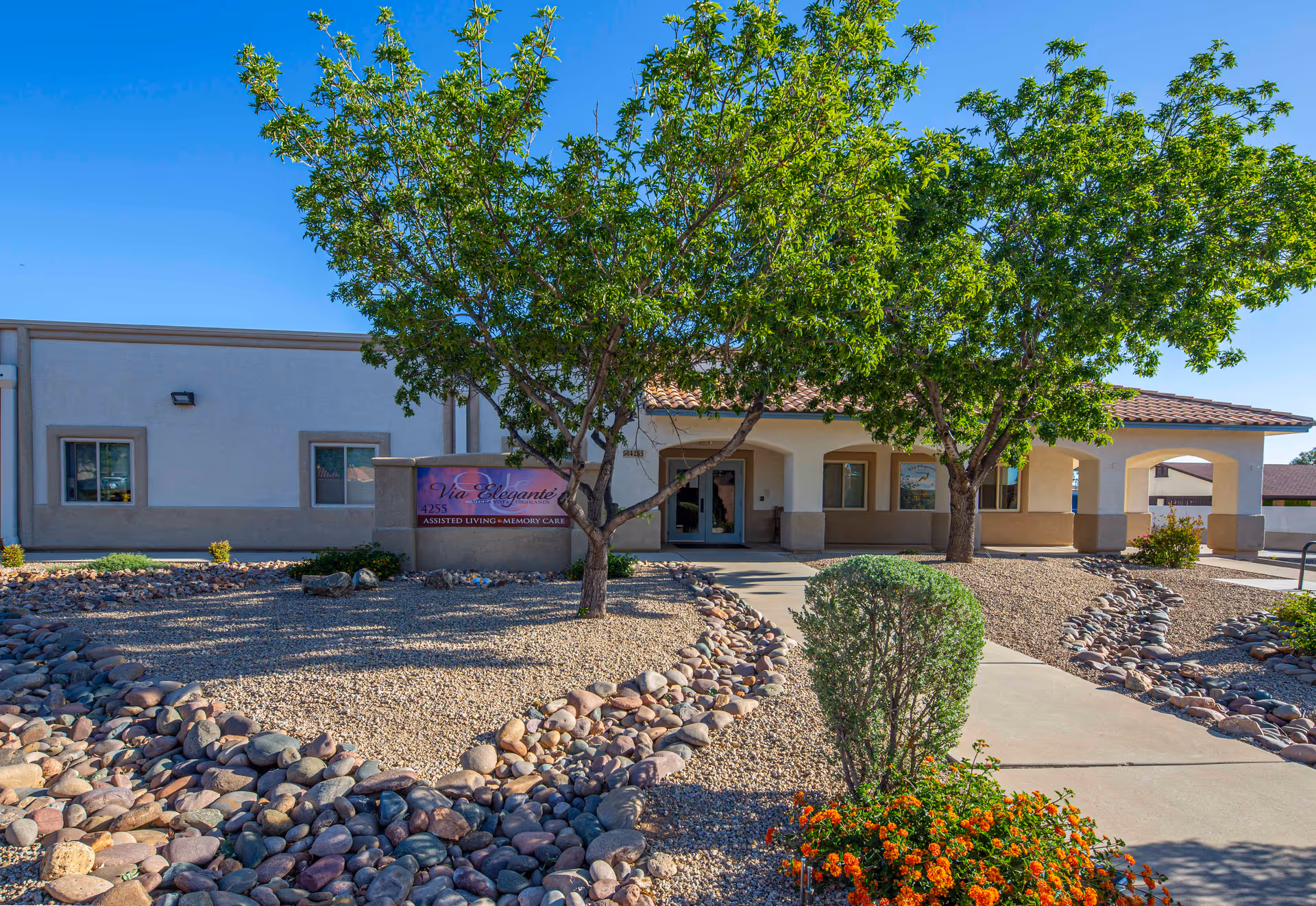 Exterior view of Via Elegante Assisted Living Sierra Vista Highlands facility showing a single-story building with a covered entrance, surrounded by desert landscaping with rocks, bushes, trees, and flowers under a clear blue sky.