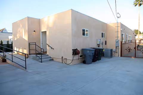Beige stucco senior facility exterior with stairs, ramp, trash bins, and a fenced gated area.