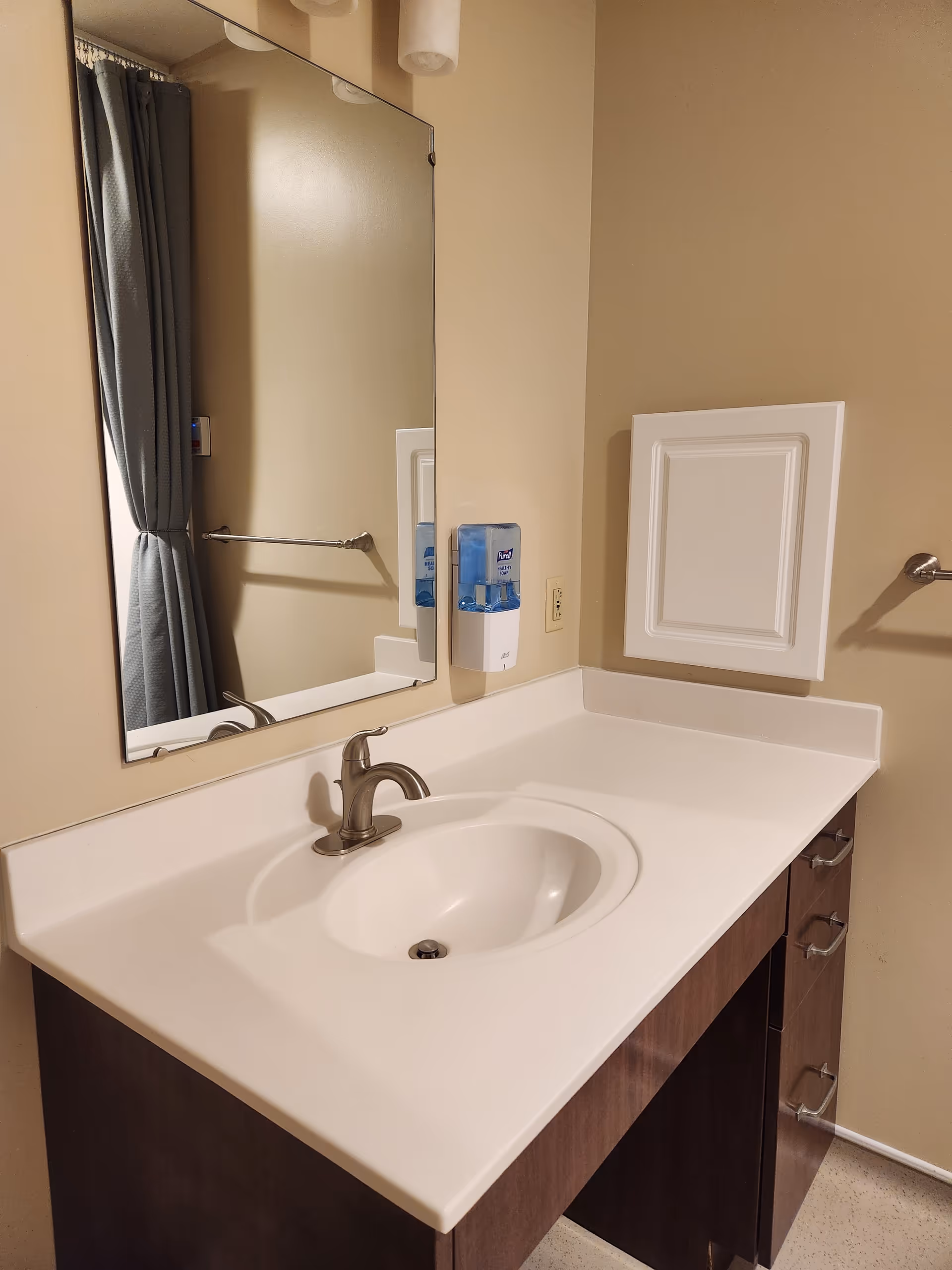 Bathroom vanity with sink, mirror, soap dispenser, and cabinet in a senior living facility.