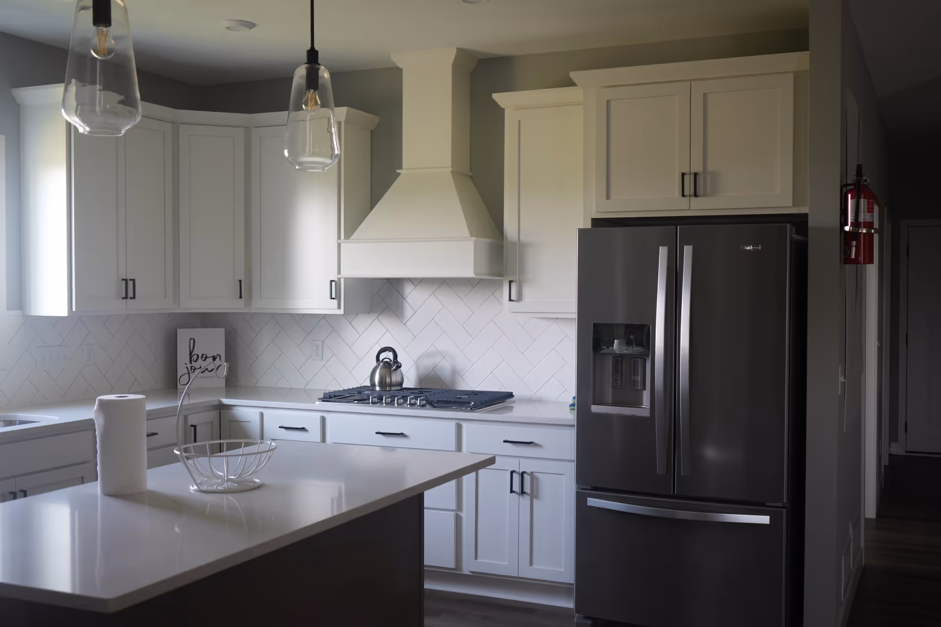 Bright modern white kitchen with island, stainless fridge, and pendant lighting.