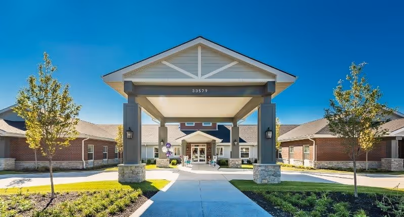 Front exterior view of Provision Living at Fenton facility with a covered entrance supported by stone and wooden pillars, a paved walkway leading to the entrance, landscaped greenery, and clear blue sky.