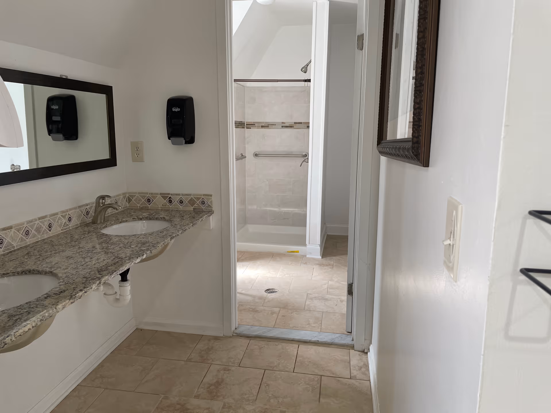 Dual-sink bathroom vanity with granite countertop, mirrors, and a tiled shower in background.