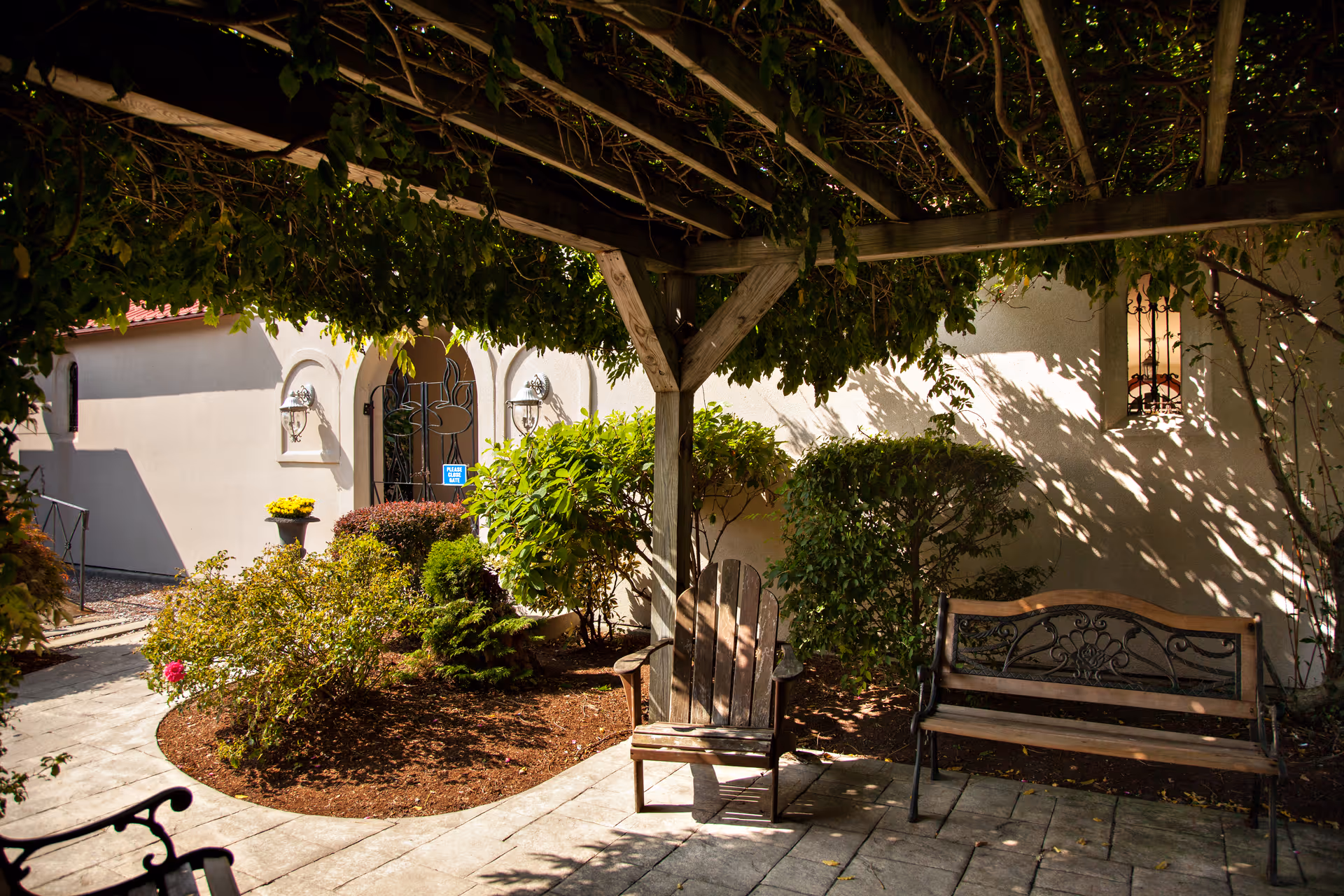 Shaded outdoor courtyard with bench, wooden chair, and surrounding greenery near the facility entrance.