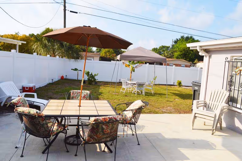 Outdoor patio area with dining table, chairs, umbrellas, and a fenced grassy yard.