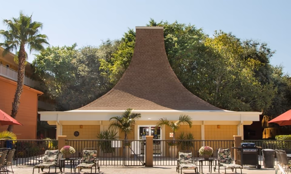 Outdoor patio area at Scottish Towers II Apartments featuring a unique building with a tall, curved roof, surrounded by trees. The patio has several lounge chairs with floral cushions, small tables with flower pots, and red umbrellas providing shade.