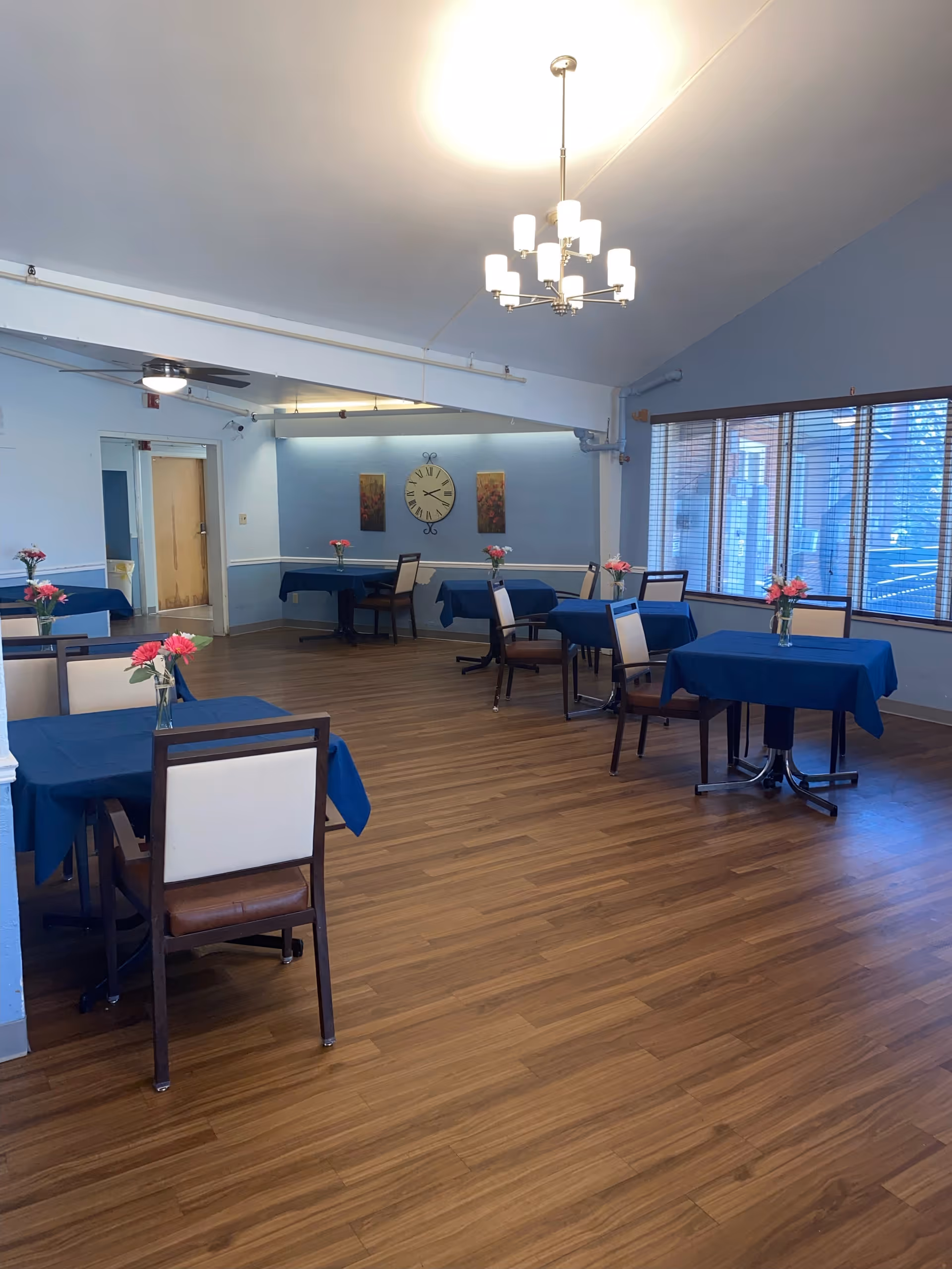 A dining room with several tables covered in blue tablecloths, each adorned with a vase of pink flowers. The room has wooden flooring, light blue walls, a large clock on the far wall, and large windows with blinds letting in natural light. A modern chandelier hangs from the ceiling.