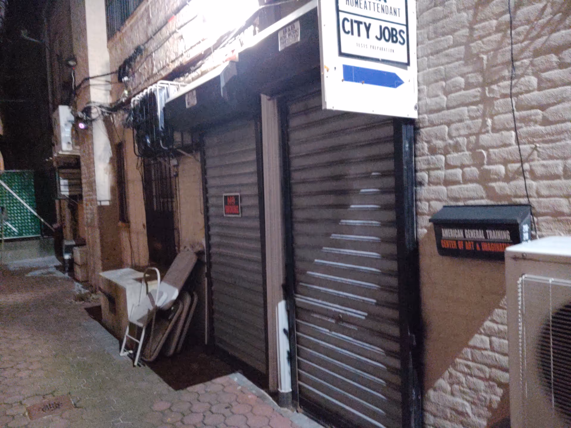 Narrow alley with closed storefront shutters and a sign for American General Training on brick wall.