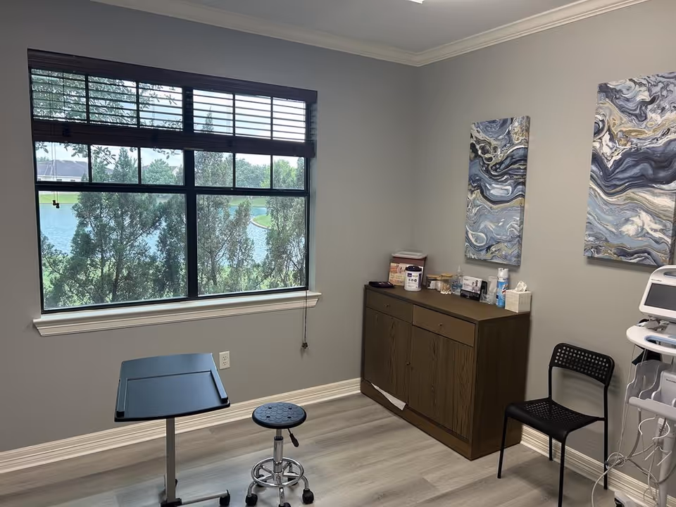 Treatment room with window view of trees and water, a medical cabinet, stool, and chair.