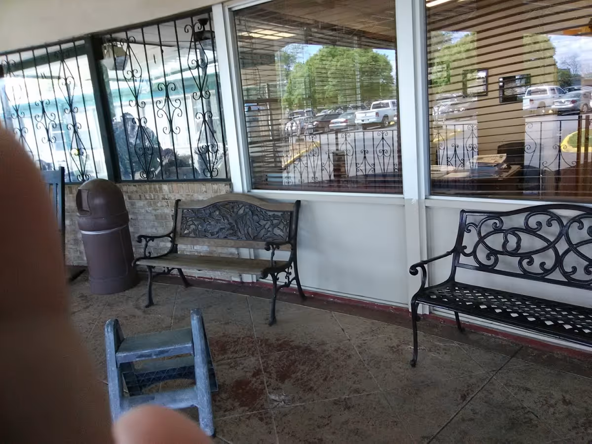Covered entrance/porch area with decorative metal benches, a trash can, and windows reflecting a parking lot.