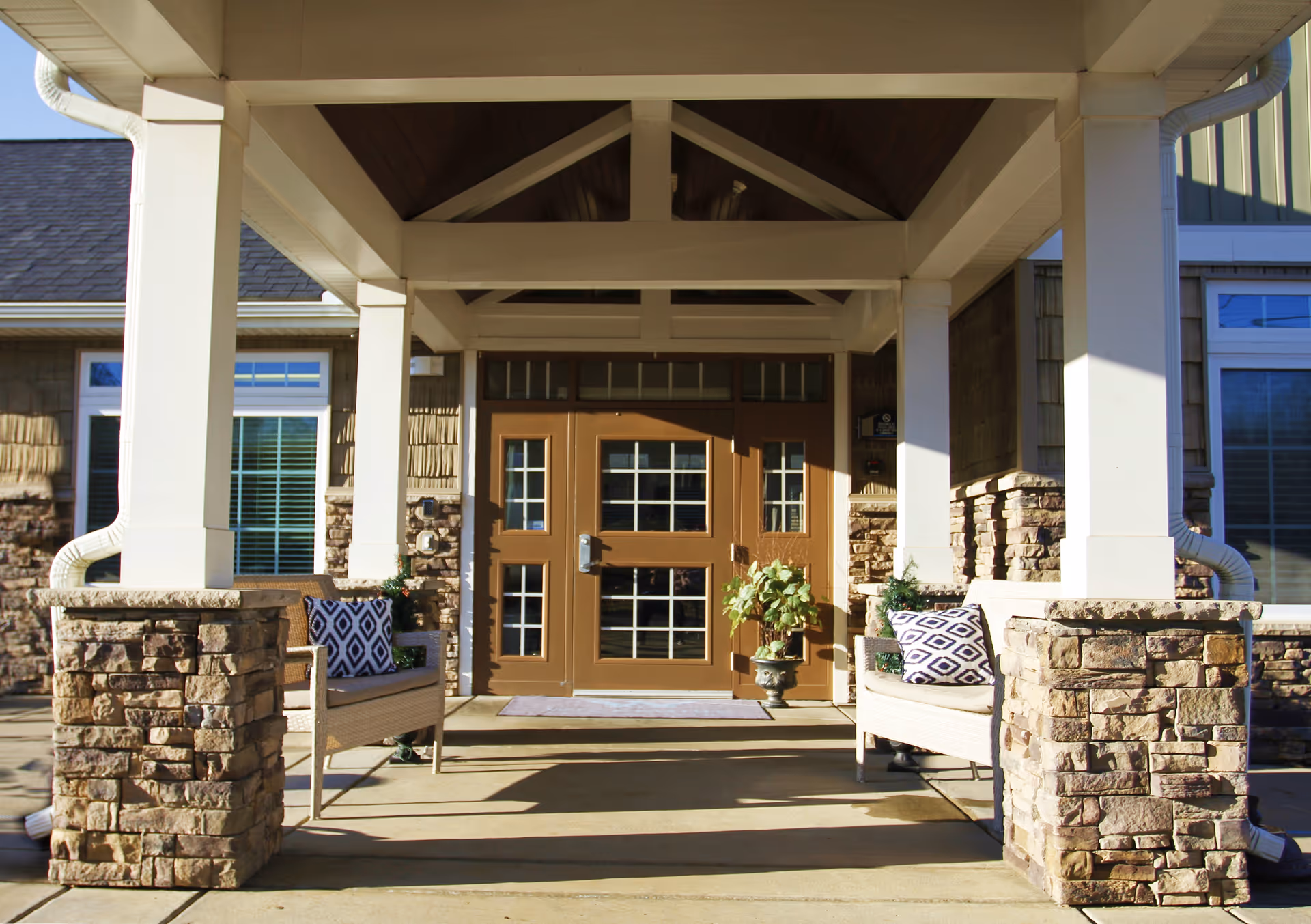 Covered building entrance with double brown doors, stone pillars, and benches with patterned cushions.