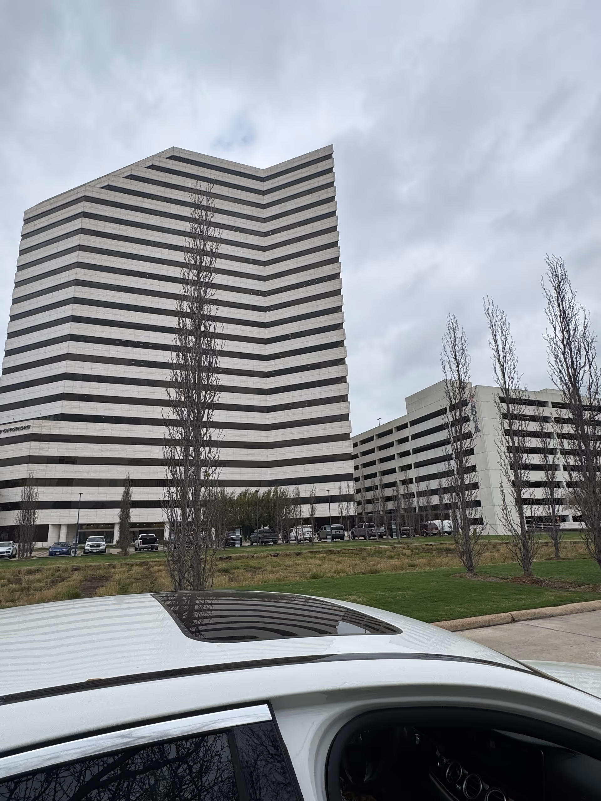 Tall striped modern building with a parking lot and leafless trees viewed from a car.