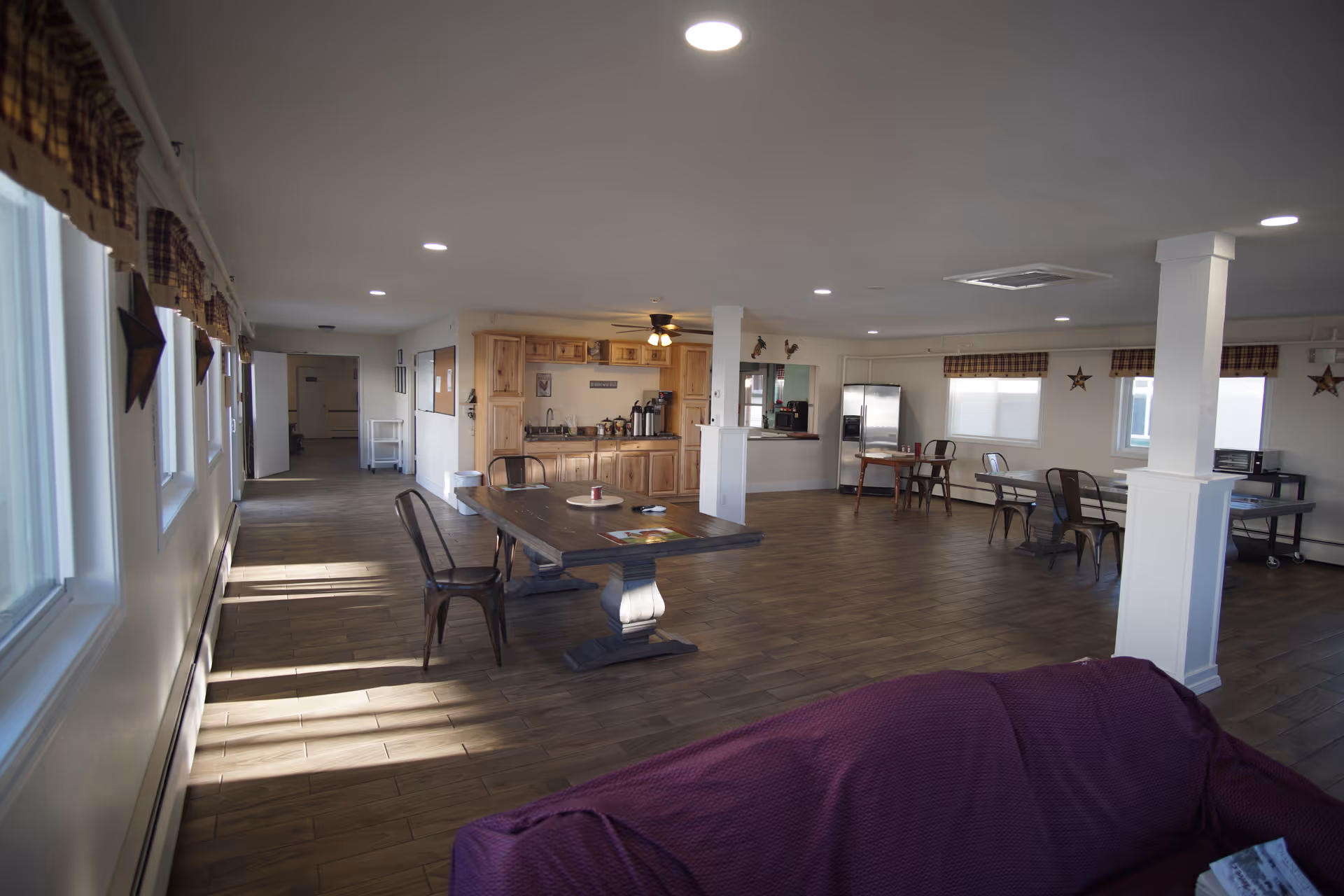 A spacious common area in an assisted living facility with wooden flooring, several tables and chairs, a kitchenette with wooden cabinets, a refrigerator, and windows with plaid valances. The room is well-lit with recessed ceiling lights and has a purple-covered couch in the foreground.