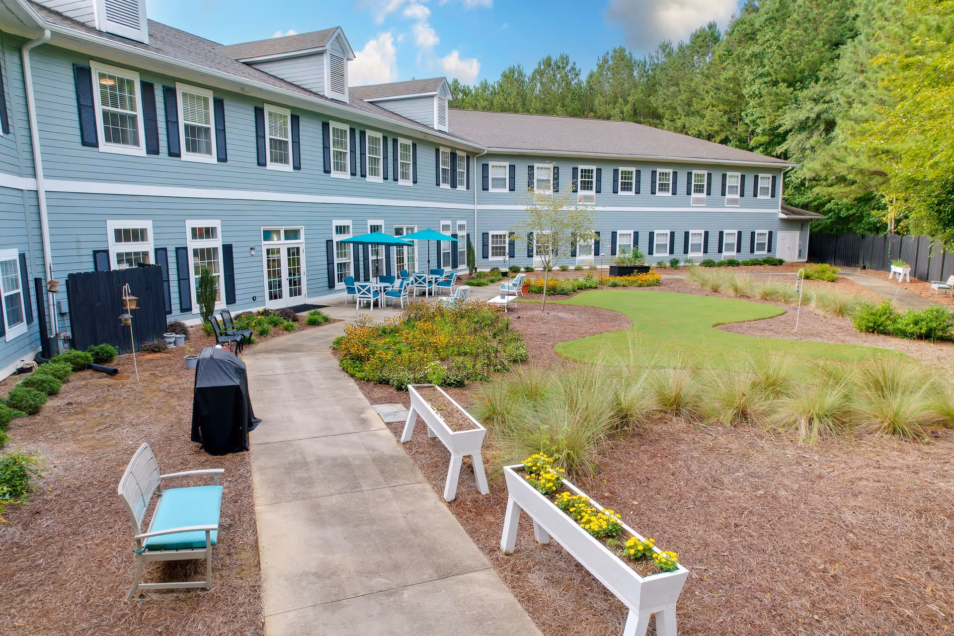 Outdoor courtyard area of a senior living facility with a two-story blue building in the background. The courtyard features a paved walkway, benches with cushions, flower planters, a putting green, patio tables with umbrellas, and various plants and shrubs. Trees and a clear blue sky are visible in the background.