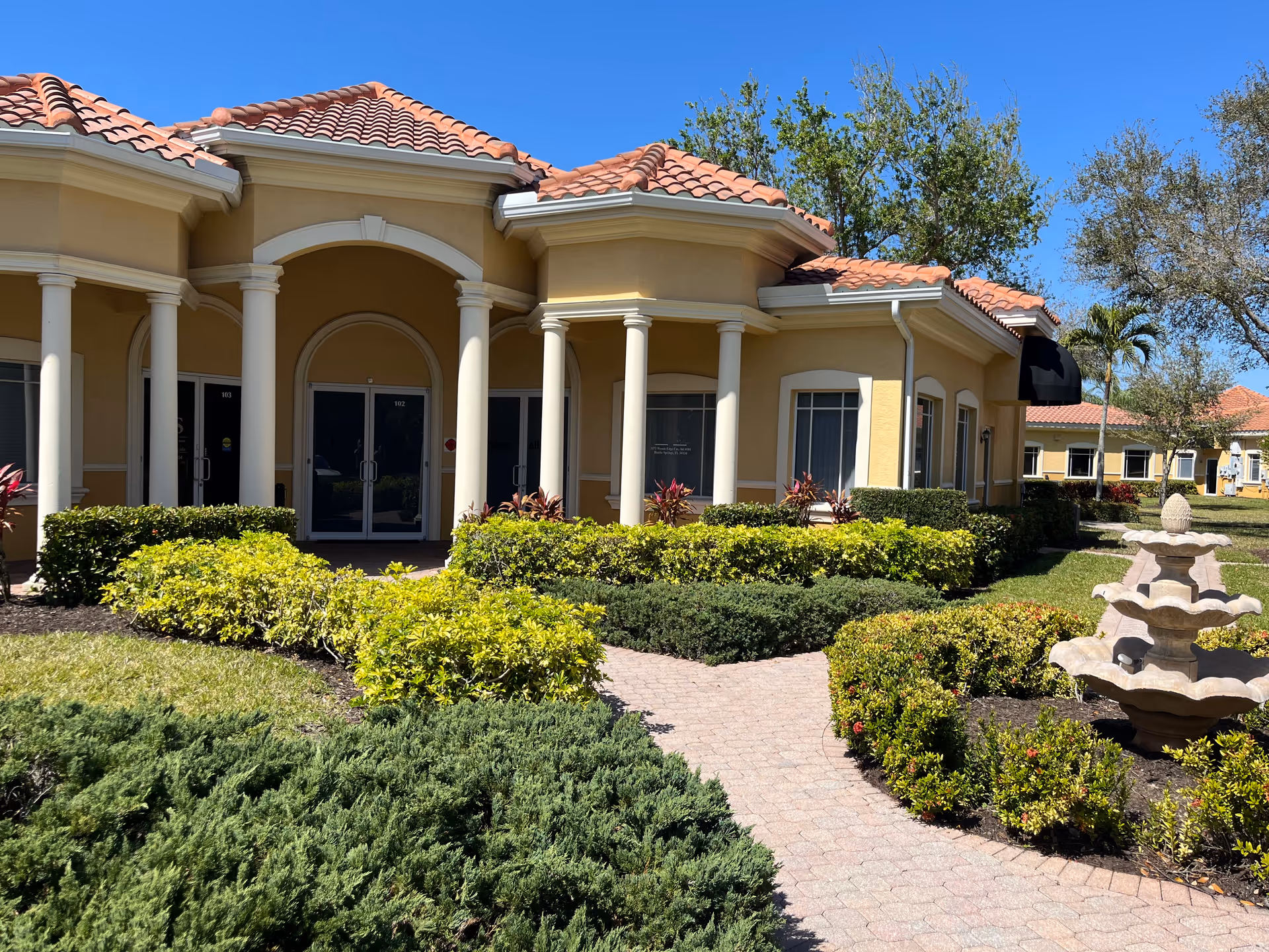 Sunlit beige senior living building with tiled roof, columns, and landscaped courtyard.
