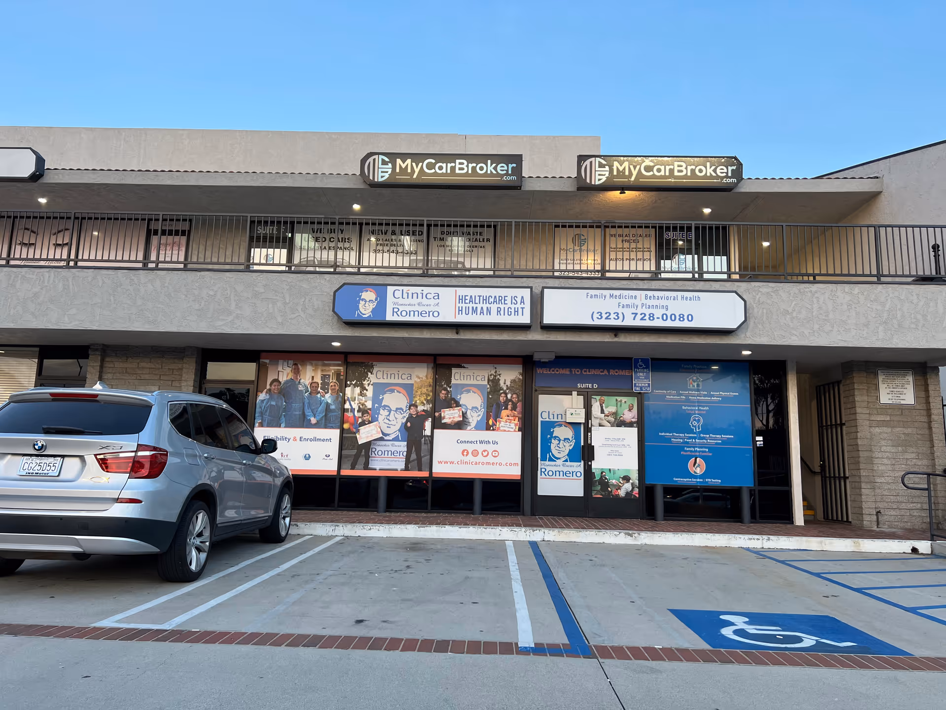 Exterior view of Clinica Monsenor Oscar A. Romero storefront with posters and a parked car.