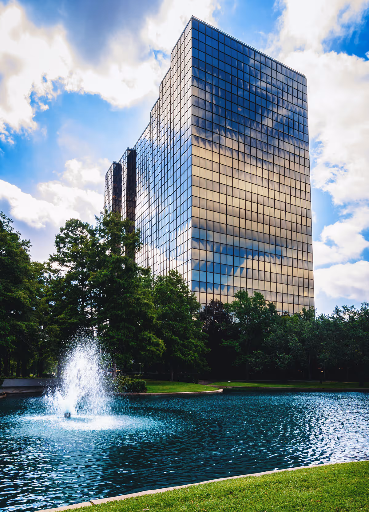 Tall glass office building reflects the sky beside a fountain and surrounding trees.