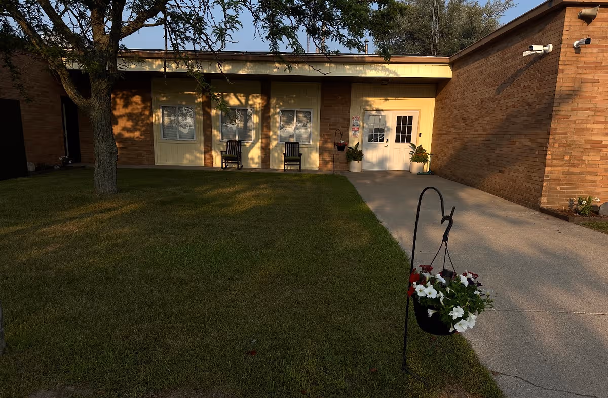 Brick senior living facility exterior with front entrance, benches, hanging baskets, and a grassy lawn.
