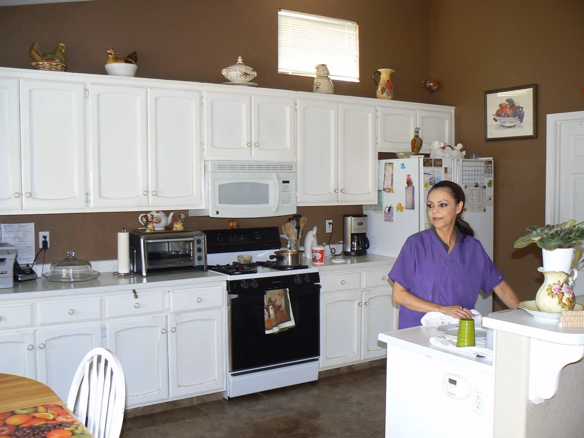 Caregiver in purple scrubs stands in a white kitchen with cabinets and appliances.
