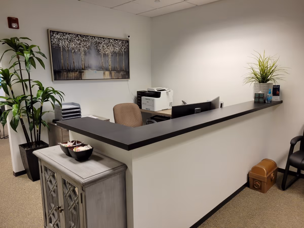 Reception desk area with a printer, computer screens, plants, and wall art.