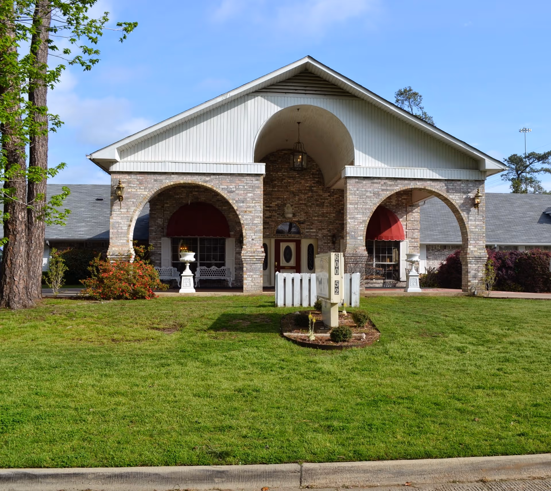 Front exterior view of Heritage Plaza Nursing Center featuring a brick building with two large arches, red awnings over windows, white benches, decorative urns, and a well-maintained green lawn in front.