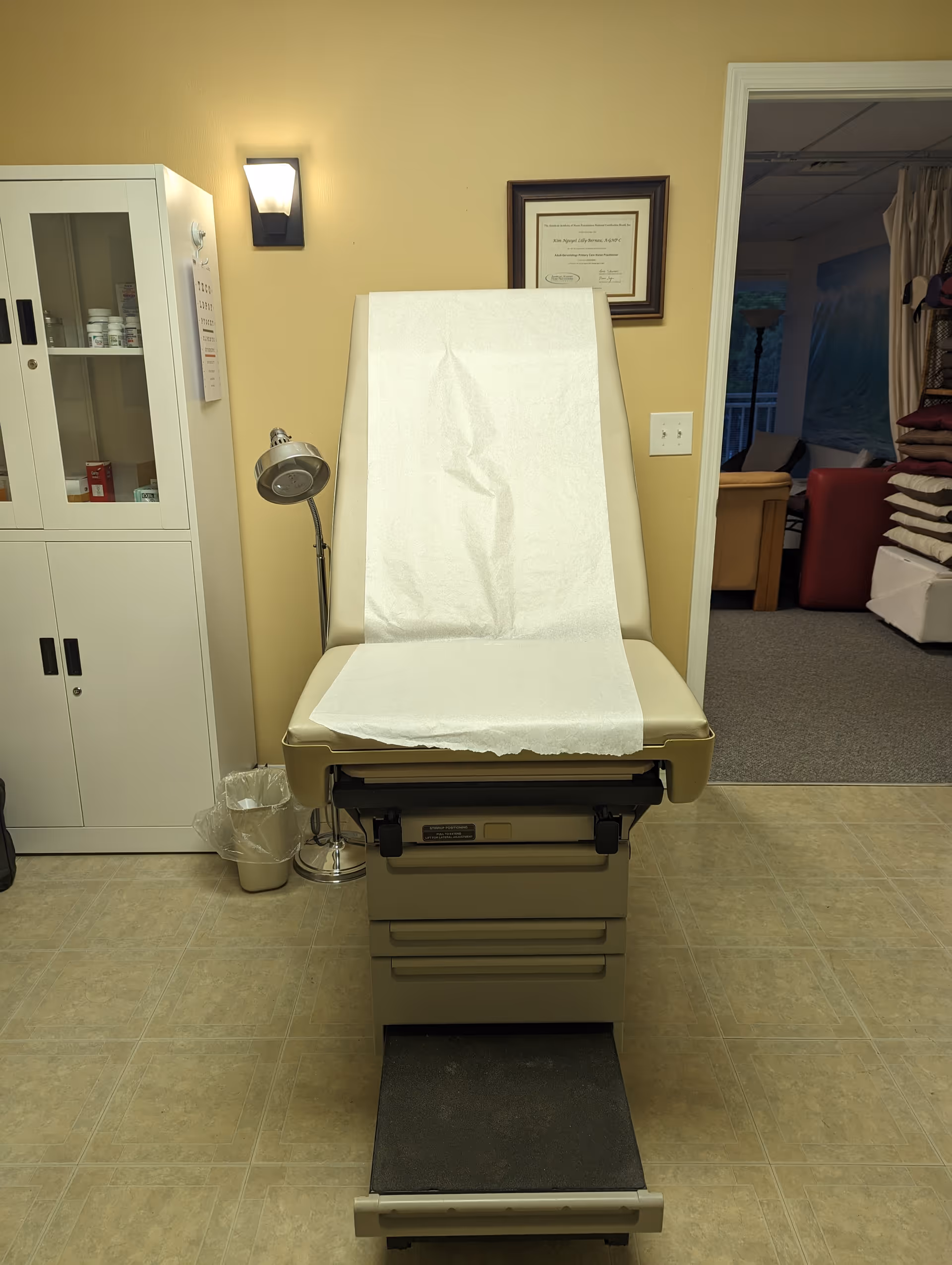 Medical examination chair in a clinic room with cabinets, lamp, and framed certificate.