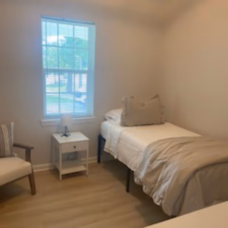 Small furnished bedroom with a bed, nightstand, lamp, chair, and window view.