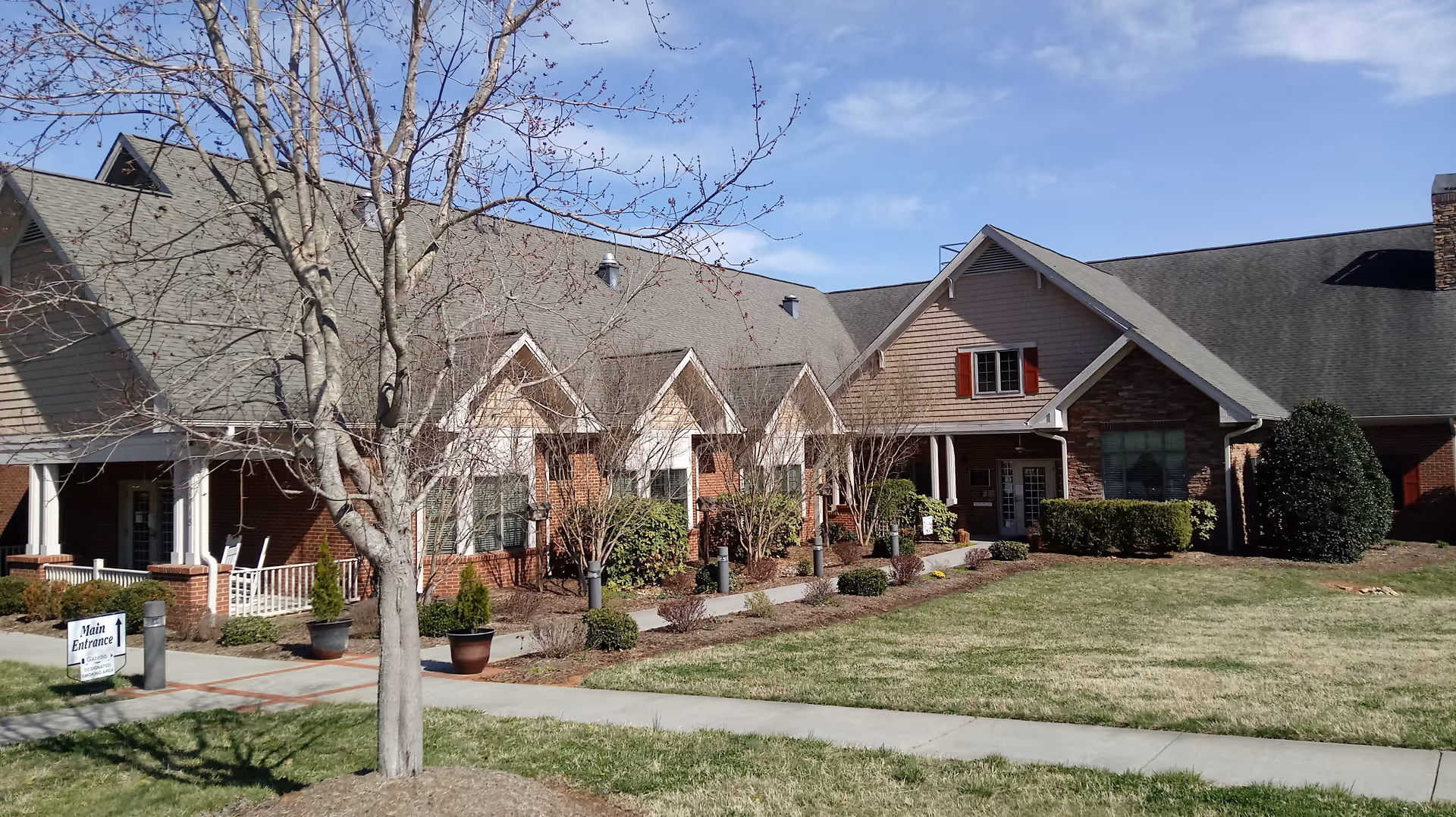 A senior living facility exterior with brick and beige siding, manicured shrubs, and a sign.