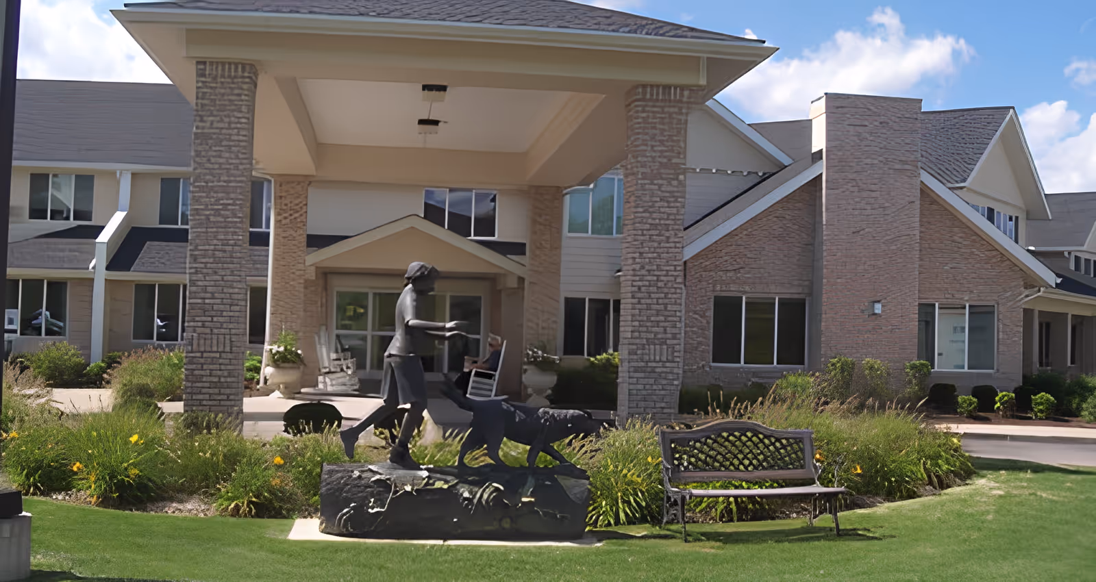 Front entrance of a senior living building with a covered porte-cochere, a statue of a person walking a dog, a bench, and landscaped lawn.