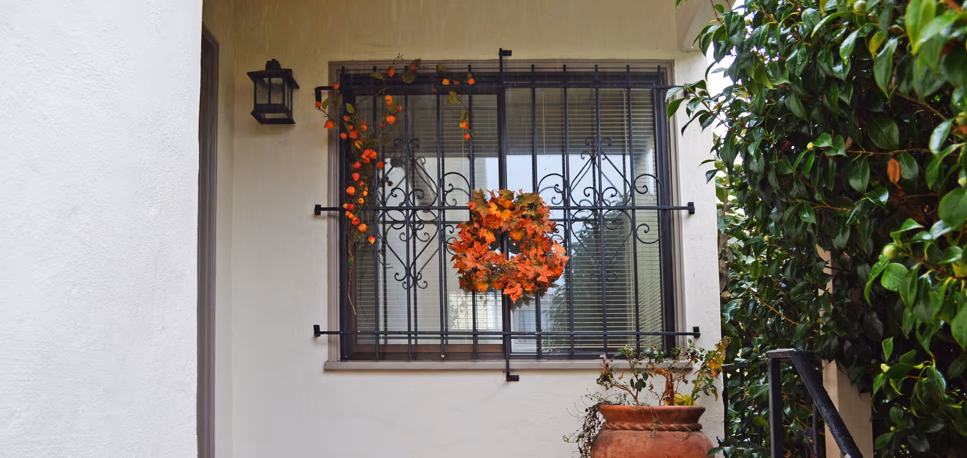 Exterior window with decorative wrought-iron bars, an autumn wreath, potted plant and surrounding greenery.