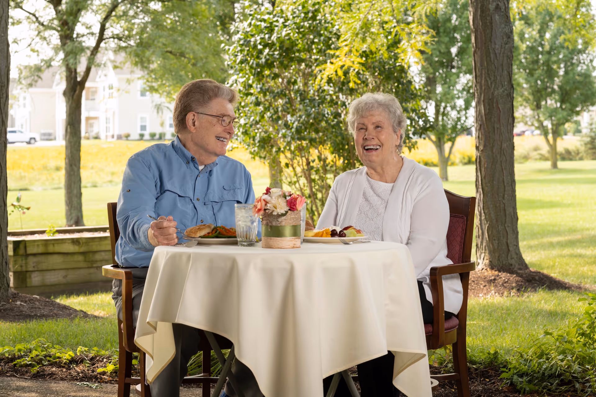An elderly man and woman sitting at a round table with a white tablecloth outdoors, enjoying a meal together. They are smiling and appear to be having a pleasant conversation. The background shows green grass, trees, and a residential building in the distance.