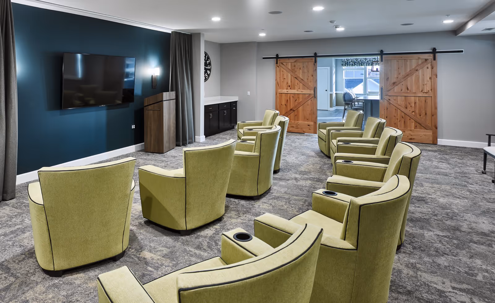 Bright media lounge with rows of green armchairs facing a wall-mounted TV and wooden sliding barn doors.