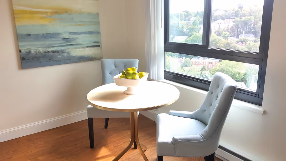 Small dining nook with a round table, two upholstered chairs, a bowl of green apples, wall art, and a large window with a view.