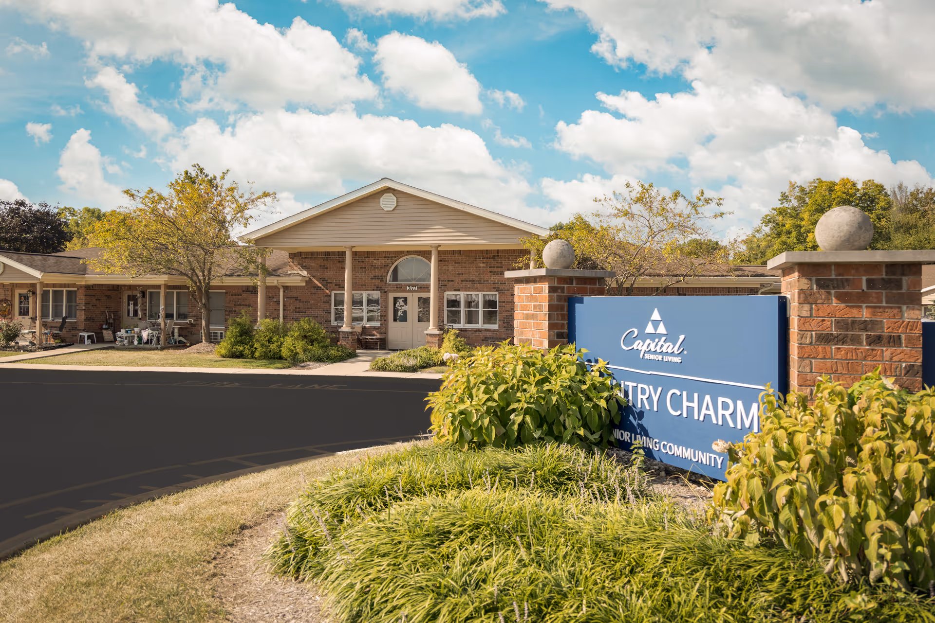 Exterior view of Country Charm senior living facility with brick building, blue sign, and landscaped entrance.