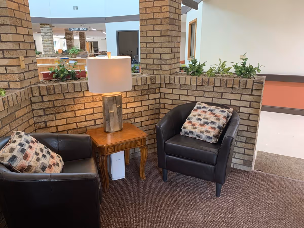 Two black leather armchairs with patterned pillows flank a wooden side table and lamp in a brick-walled seating nook inside an assisted living facility.
