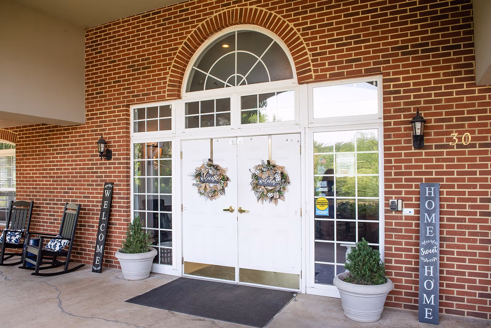 Entrance to a brick building with double white doors decorated with wreaths. Two potted plants flank the doors, and there are rocking chairs with cushions on the left side. Signs next to the doors read 'WELCOME' and 'HOME Sweet HOME'. The building number 30 is visible on the right side.